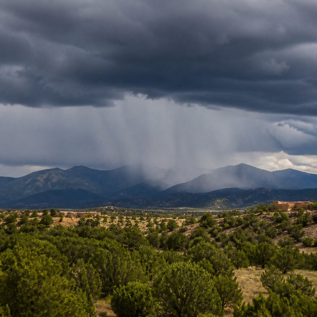 A dark monsoon storm cell standing over the Sangre de Cristo mountains on a summer afternoon, grey columns of rain falling beneath it while the ground in front stays sunlit