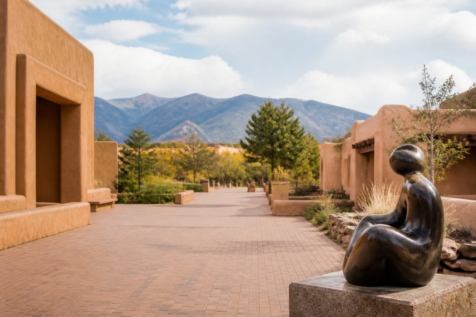 The paved entrance plaza on Museum Hill, low adobe museum buildings on either side, a bronze sculpture in the foreground and the mountains beyond