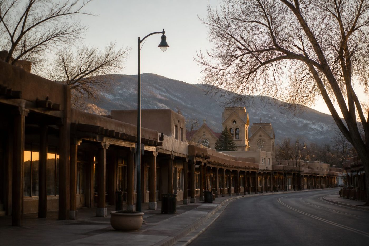 Early morning on the Santa Fe Plaza: low adobe buildings with a covered walkway, bare cottonwood branches and the Sangre de Cristo foothills behind